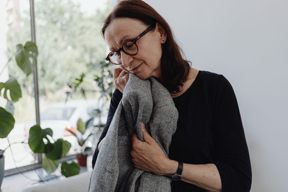 A senior woman wearing glasses stands indoors holding a gray blanket, deep in thought.