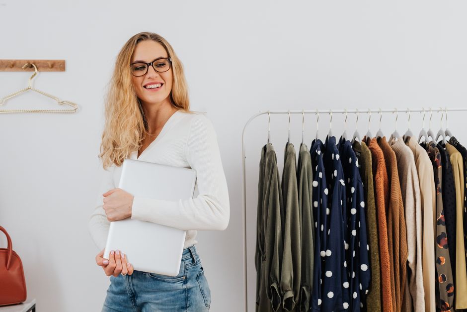 Smiling woman with glasses holding laptop next to stylish wardrobe indoors.