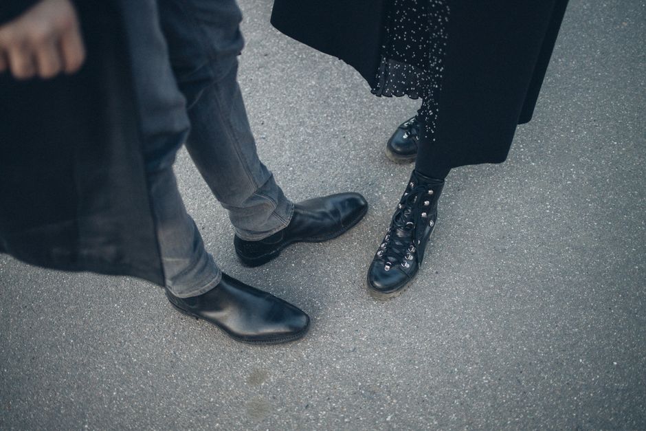 A fashionable couple shows off their black leather footwear while standing together on a city street.