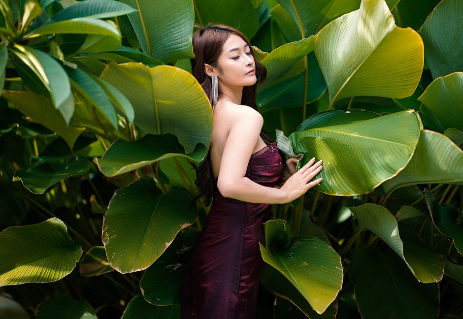 Woman in a burgundy dress posing gracefully among tropical leaves.