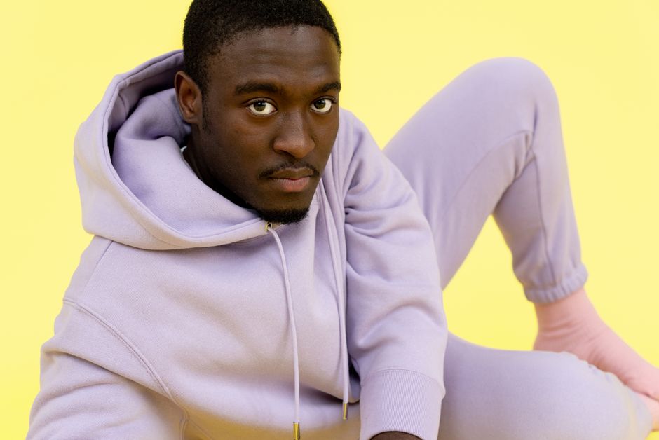 Portrait of a young man in a stylish lavender hoodie seated against a bright yellow backdrop.