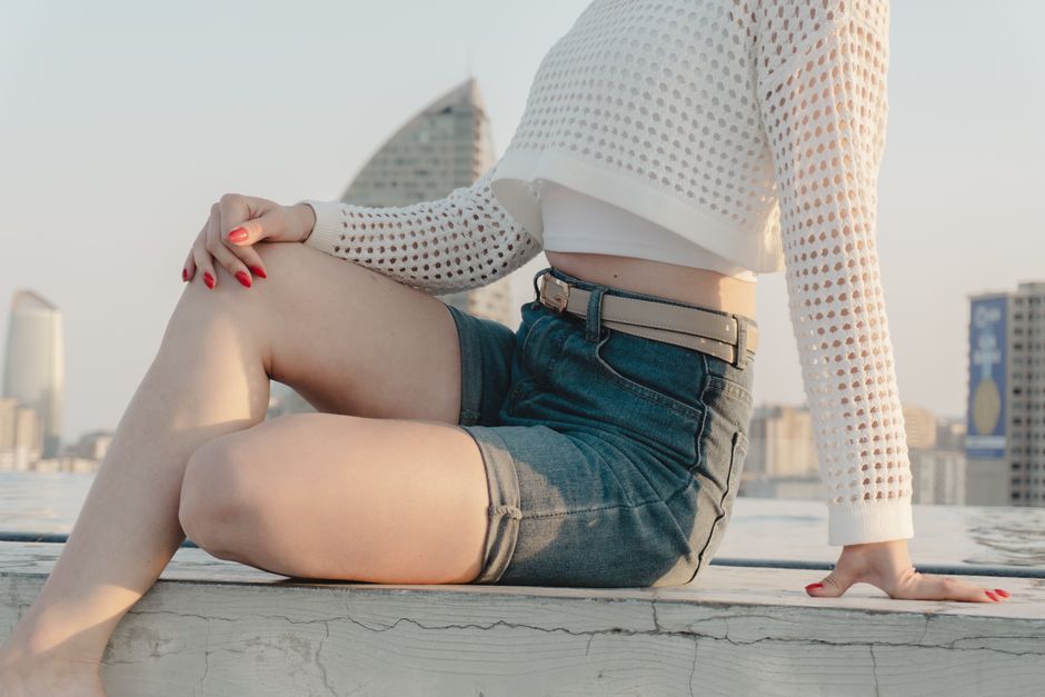 Woman in shorts and sweater poses on a rooftop with cityscape in the background.
