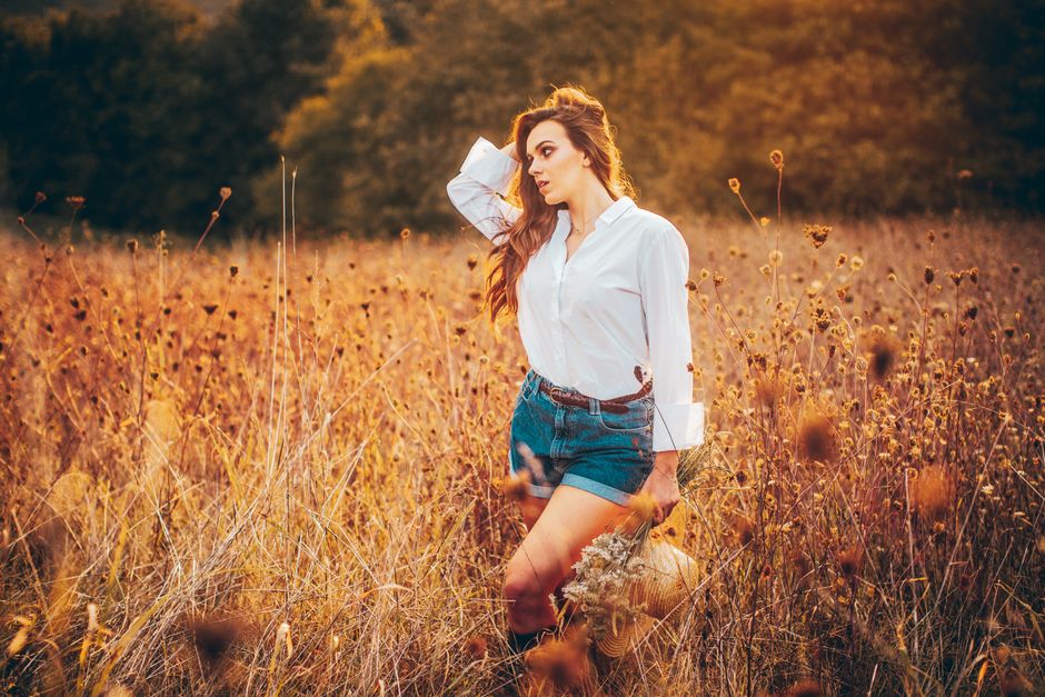 A woman in denim shorts and white shirt poses gracefully in a sunlit autumn field.