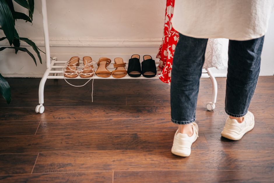 Person in casual attire browsing shoes on a rack indoors, wooden flooring visible.