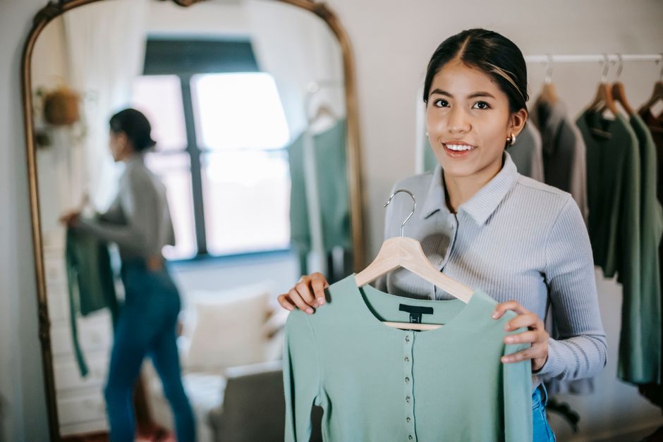 Smiling young ethnic woman in blouse and jeans standing with blue cloth on hanger at home near wardrobe and mirror while looking at camera