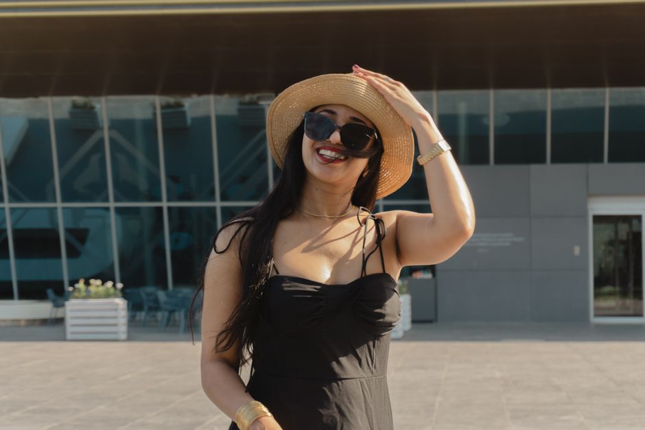 Smiling woman in black dress and straw hat enjoying a sunny day in Baku, Azerbaijan's urban landscape.