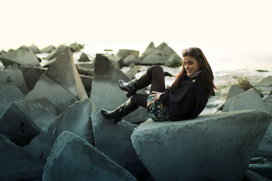 A fashionable woman in black boots posing on geometric concrete blocks near the sea.