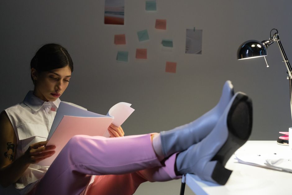 Woman in a modern office setting reading documents, feet up on desk under warm light.