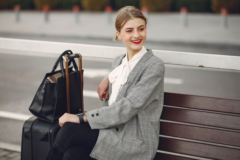 Positive female traveler in trendy clothes smiling away while waiting for train sitting on wooden bench with suitcase and bag in street