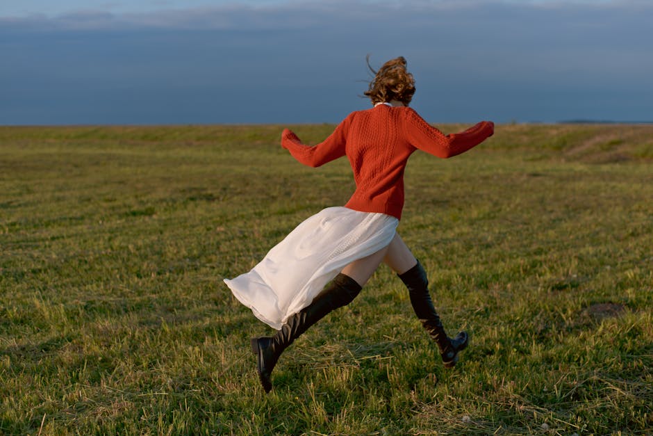 A woman in boots runs energetically across a grassy field under a clear sky, embracing freedom and nature.