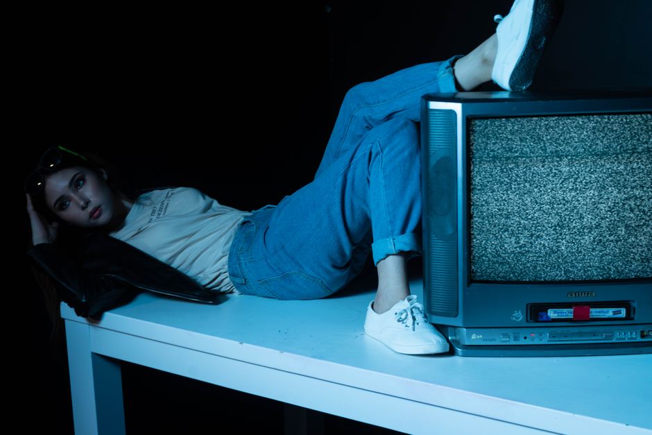 Young woman in casual attire posing with vintage television on table in dimly lit room.