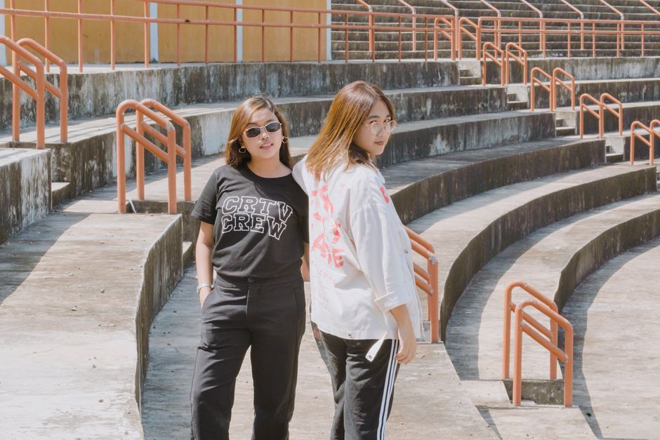 Two young women standing in an empty outdoor stadium with concrete seating and orange railings in bright sunlight.