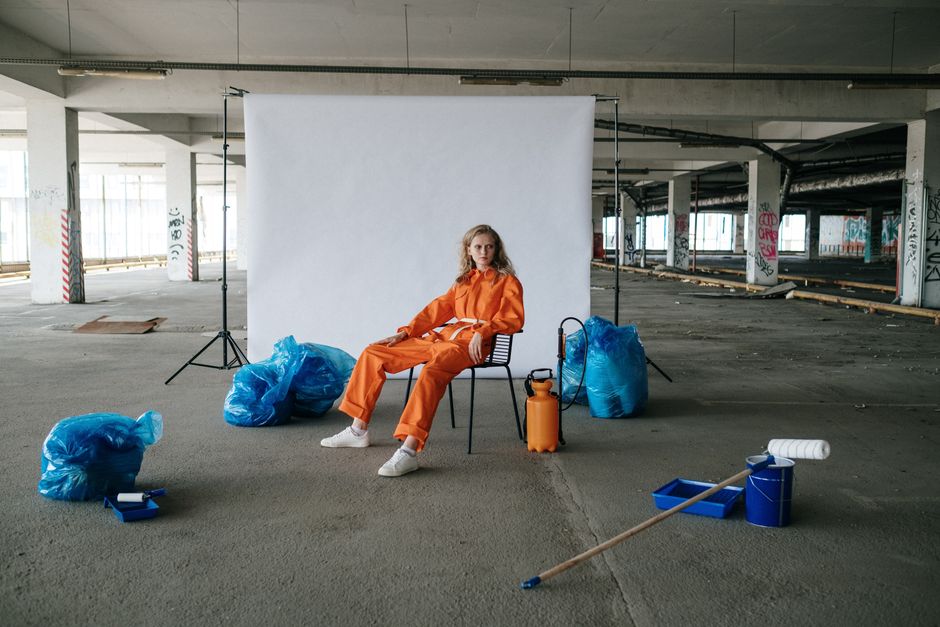 Artistic portrait of a woman in orange jumpsuit sitting in an industrial space with props.