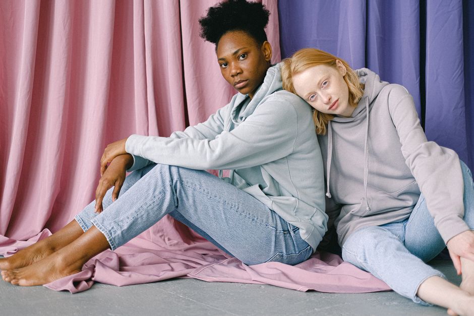 Two women sitting on the floor in jeans and hoodies, with a backdrop of pastel curtains.