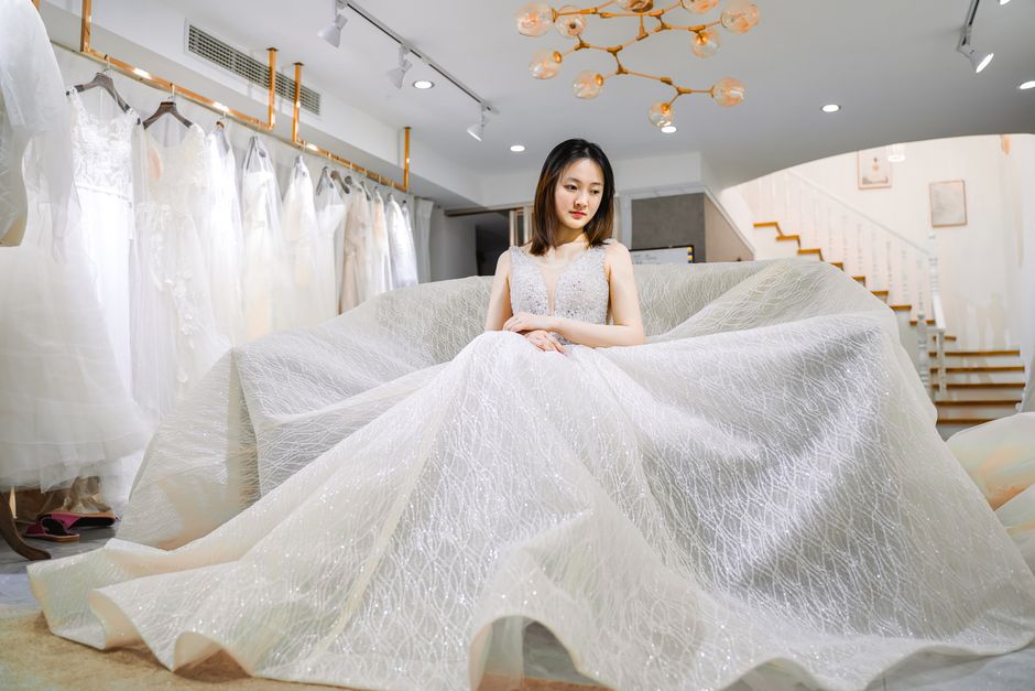 Pondering ethnic female bride in white ornamental dress looking away while sitting near collection of wedding dresses behind stairs in bridal salon with lamps on ceiling