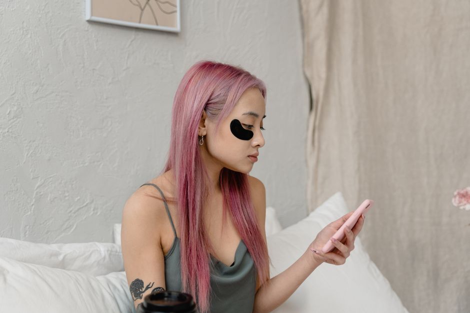 Asian woman relaxing with under-eye patches while looking at her phone indoors.