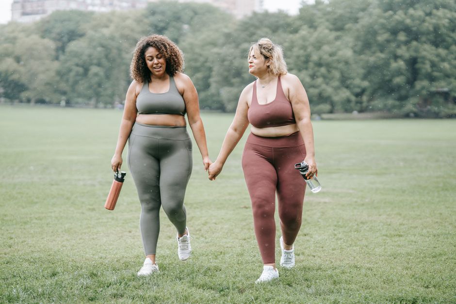 Two plus-size women in activewear holding hands while walking in the park, promoting fitness and friendship.