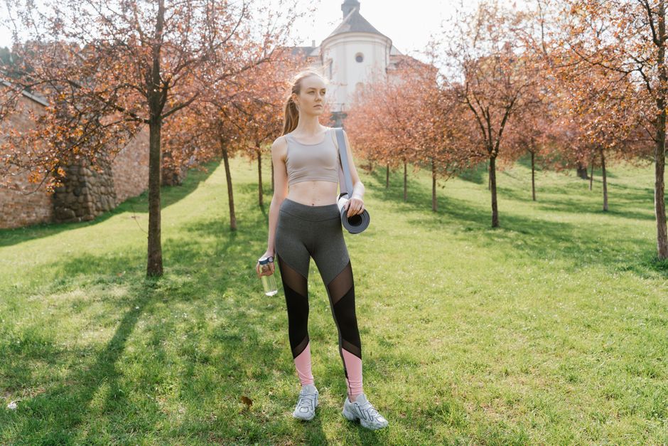 Athletic woman holding water bottle and mat, posing outdoors in sunny park.