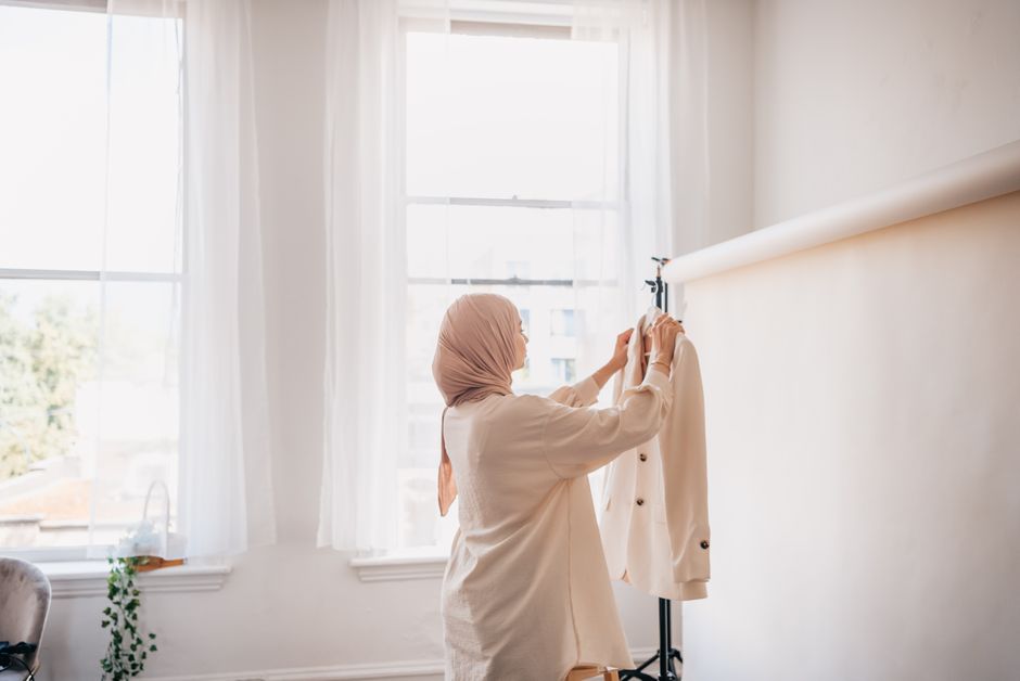 A woman in hijab holds a white blazer in a bright, airy home setting.
