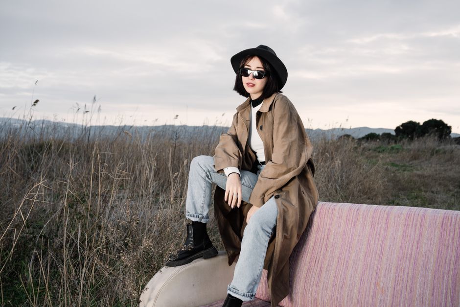Young woman in trench coat and sunglasses sitting on a couch in a field, showcasing modern fashion style.
