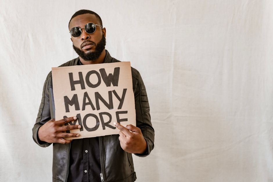 Man holding cardboard sign with 'How Many More' on a neutral background.