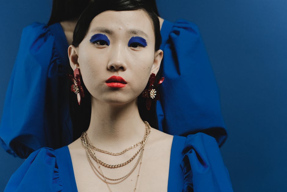 Studio portrait of an Asian woman with bold makeup, blue dress, and statement jewelry.