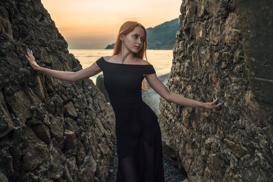 A woman in a black dress poses elegantly between rocks on a beach at sunset in Гагра.