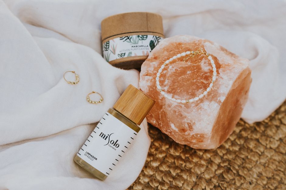A flat lay of natural skincare products, a mineral stone, and jewelry on fabric.