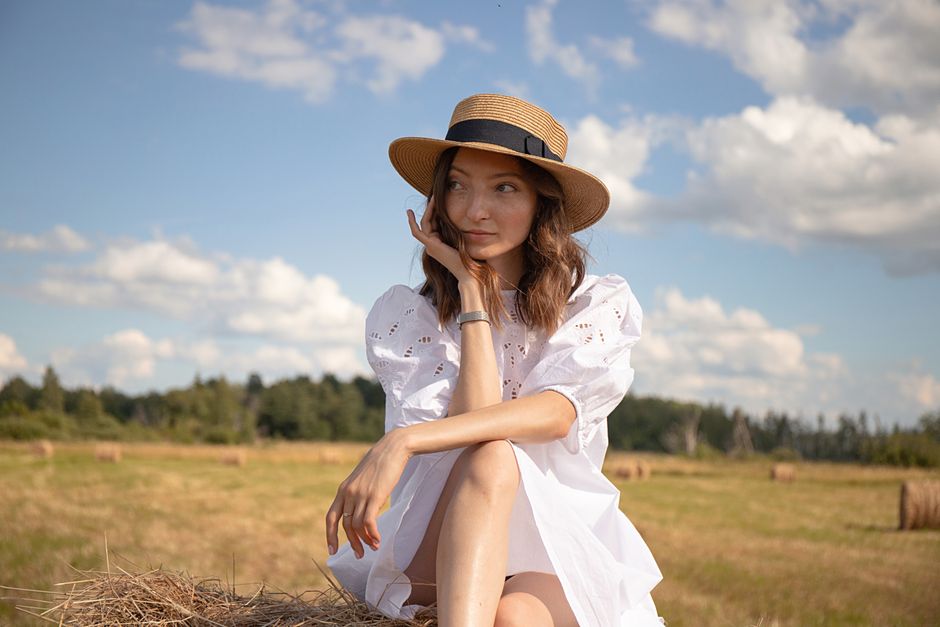 Elegant woman in white dress and straw hat posing gracefully in an open summer field with hay bales.