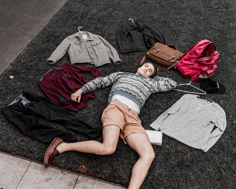 A young man lies on grass surrounded by clothes and bags in an outdoor setting.