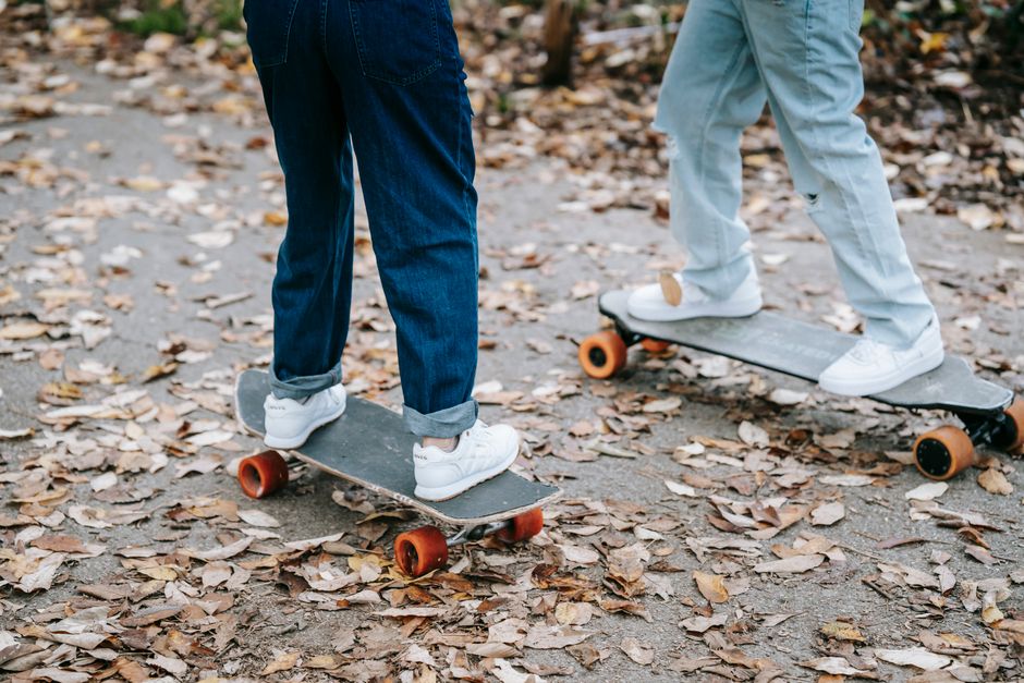 Crop legs of anonymous people in jeans and sneakers riding skateboards on asphalt walkway in park in autumn day
