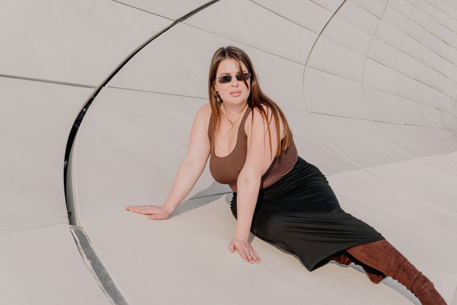 A fashionable woman wearing sunglasses poses against a contemporary architectural wall.