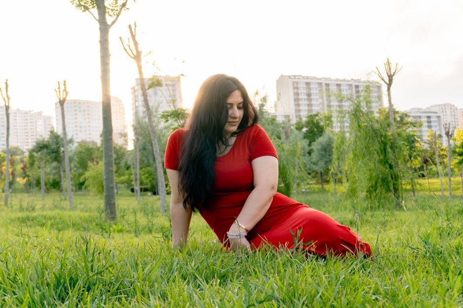 A woman in a red dress sits on grass in an urban park with buildings in the background.