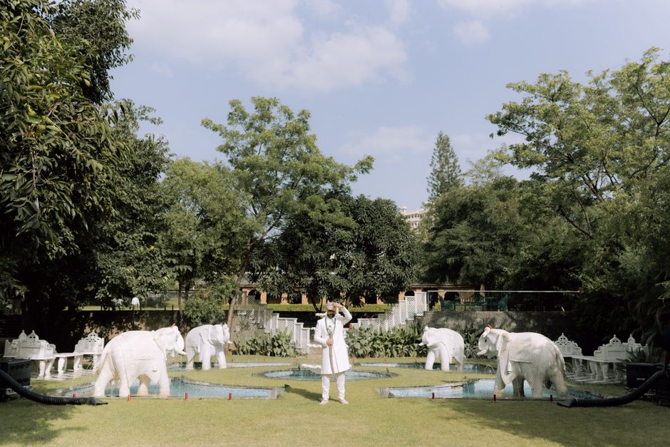 A man in traditional attire stands in a decorative garden with white elephant statues.