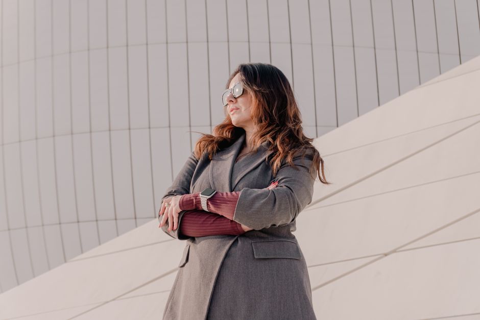 A confident woman in a grey coat and sunglasses stands against a modern architectural backdrop.