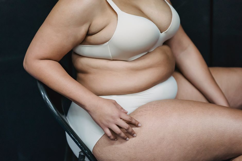 Crop anonymous plump woman in white underwear sitting on chair in light studio on dark background