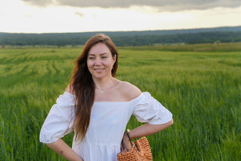 Smiling woman in a white dress standing in a lush green field, holding a woven basket under a cloudy sky.