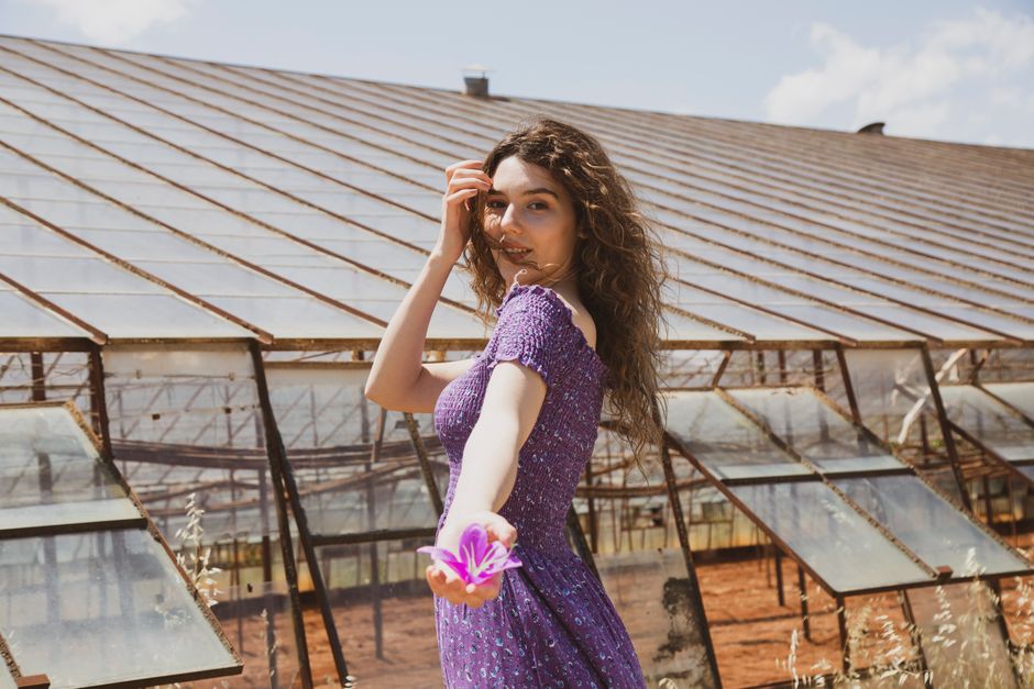 A woman in a purple dress posing with a flower in front of a greenhouse. Perfect for lifestyle or fashion themes.