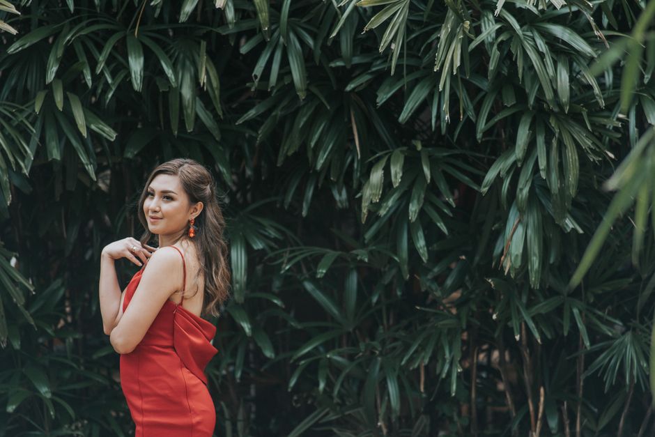 Elegant woman in a red dress posing gracefully against lush green foliage.