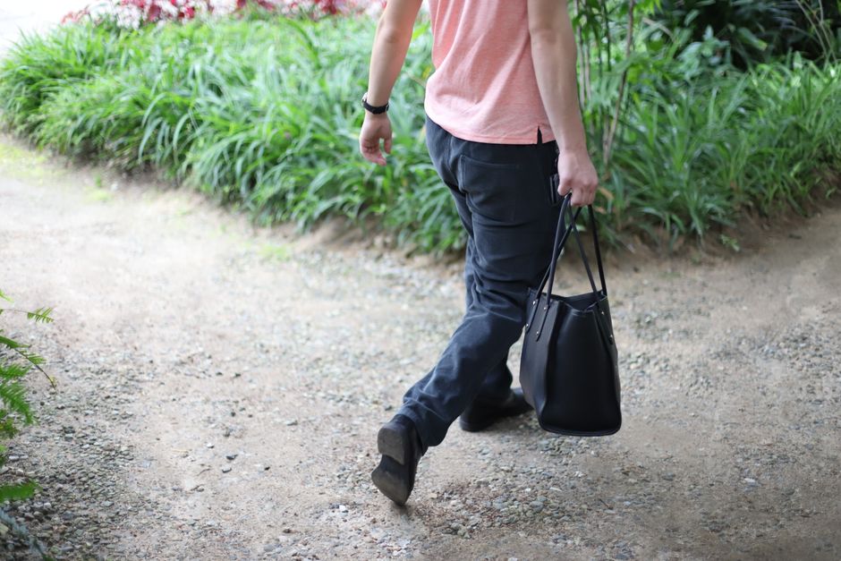 A stylish man in casual attire walking on a garden path carrying a tote bag.