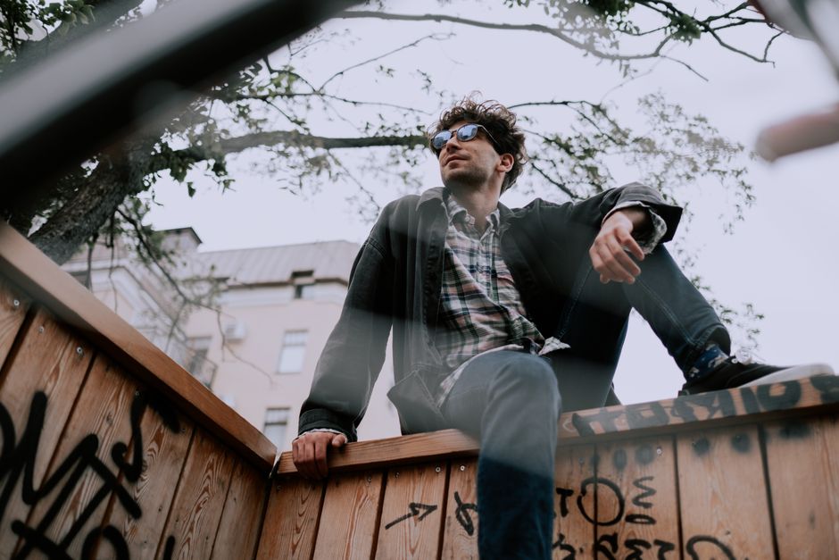 Man sitting on wooden ledge with graffiti, wearing sunglasses in an urban outdoor setting.