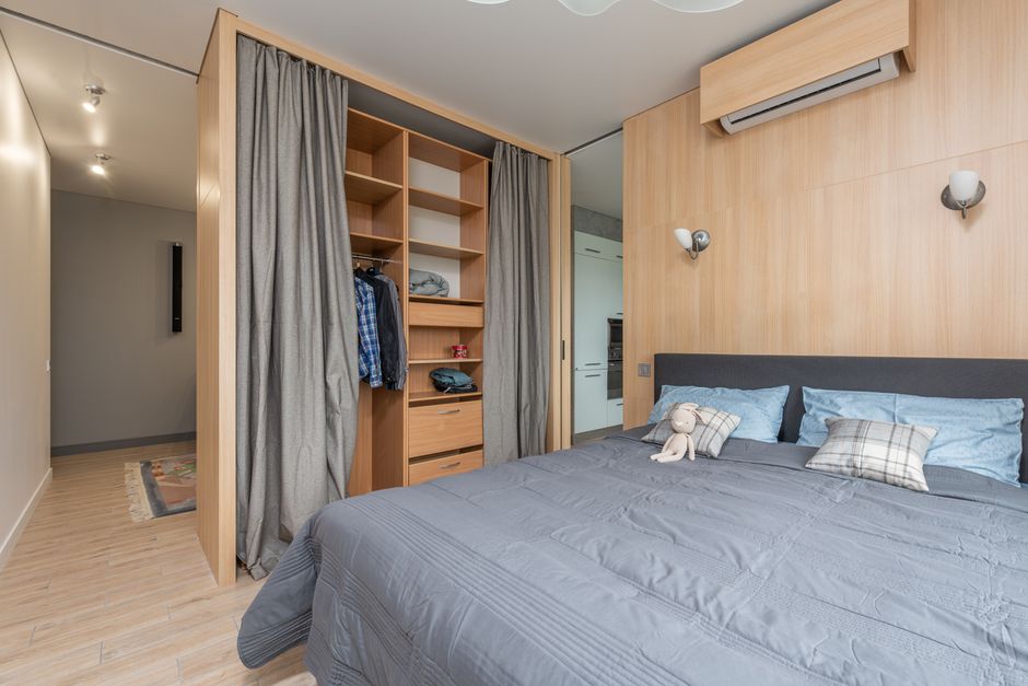 Modern room interior with bed with pillows and soft rabbit toy near wooden cupboard with curtains and shelves near clothes and entrance to kitchen in bright home