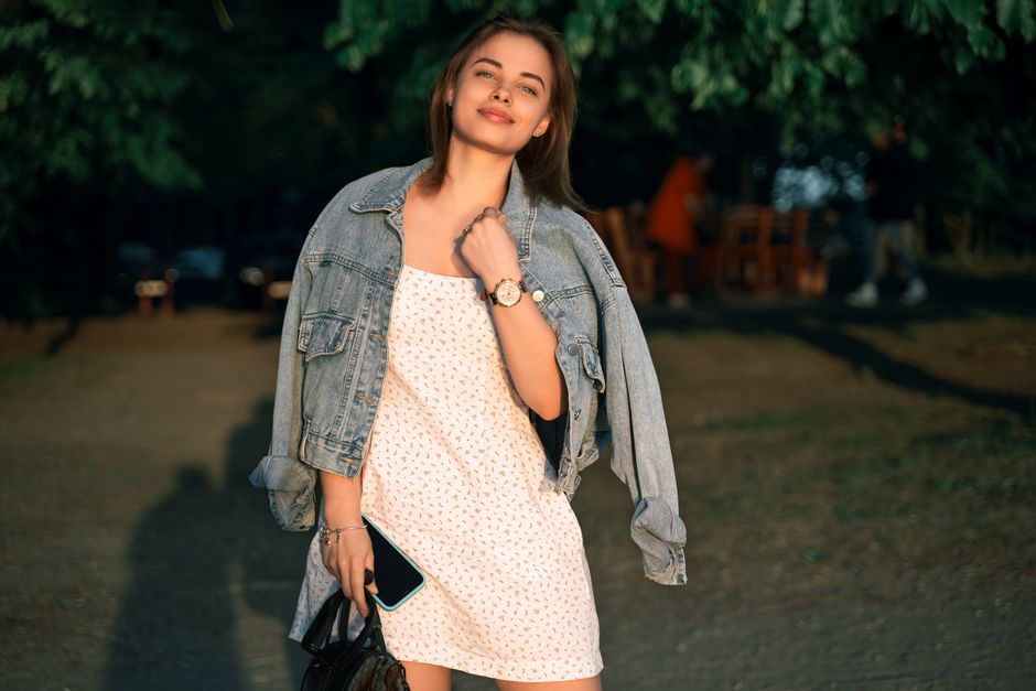 Fashionable woman smiling in a jean jacket and dress outdoors in a serene setting during sunset.