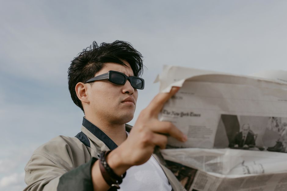 A stylish adult male in sunglasses reads a newspaper under a cloudy sky.