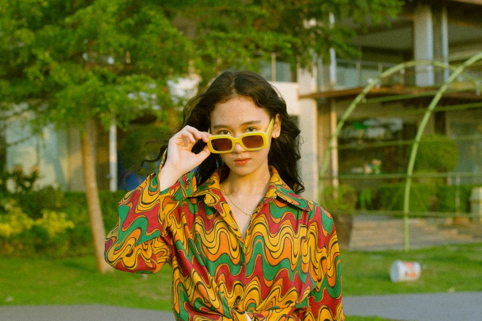 Stylish young woman posing with yellow sunglasses and colorful outfit in a sunny outdoor setting.