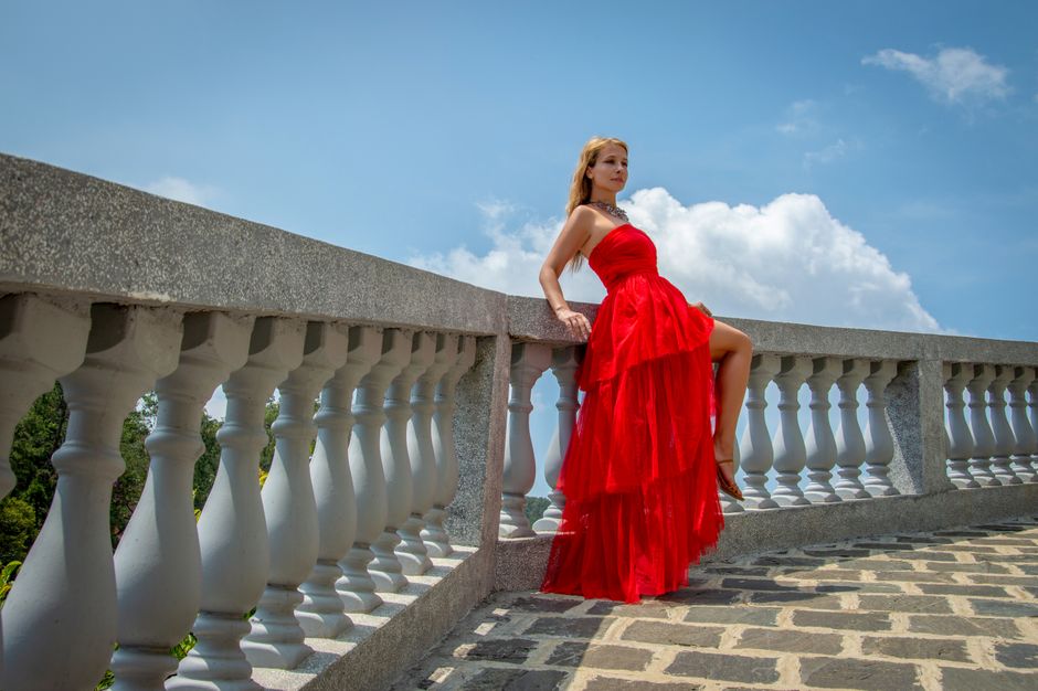 A woman in a red dress poses elegantly on a stone balcony against a clear sky.