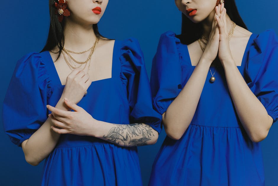 Two fashionable women in blue dresses, showcasing tattoos against a matching blue background.