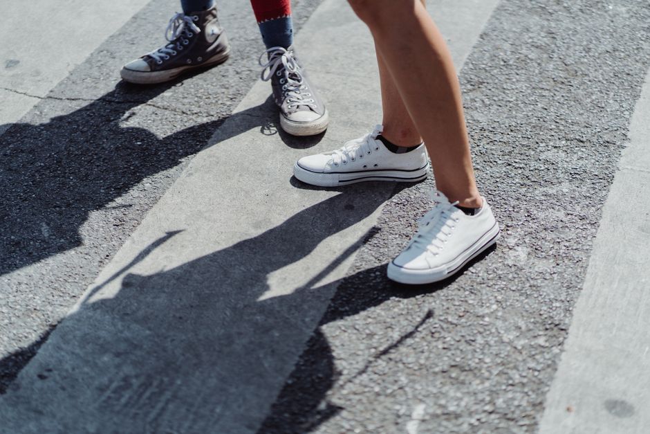 Two individuals in canvas sneakers standing on a city street crosswalk on a sunny day.