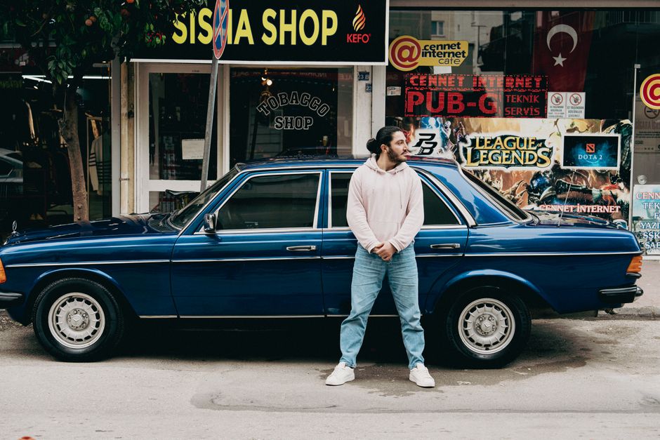 Young man in casual attire leaning on a vintage blue car on a busy urban street.