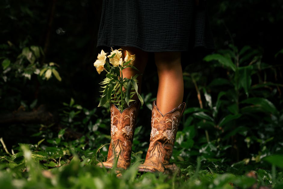 A woman stands in a forest wearing cowboy boots, holding yellow flowers.
